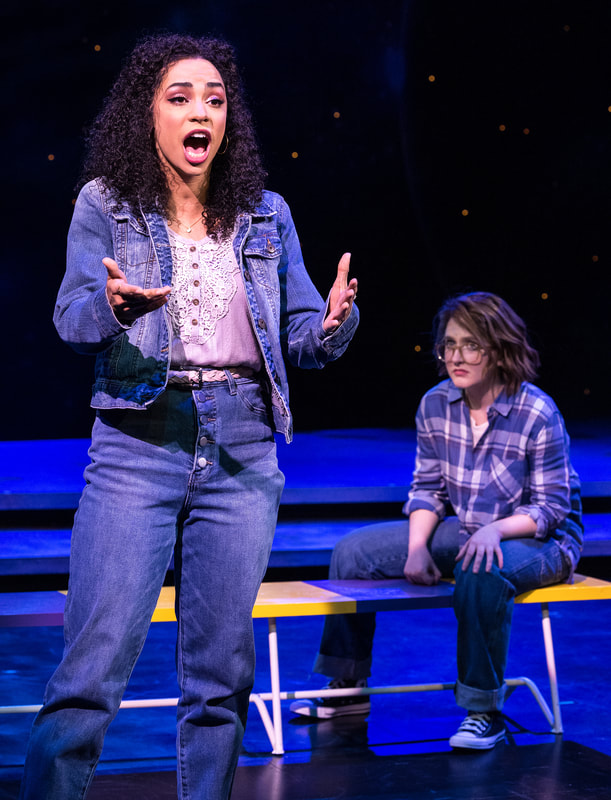 An actor in denim stands on stage holding hands out and singing, another actor sits on a bench watching.