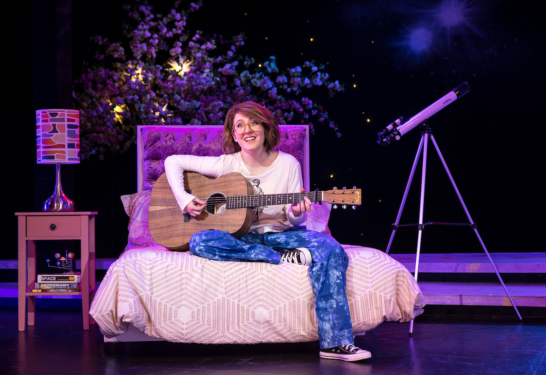 Actor sits on a bed, on stage, holding a guitar and singing. Telescope and stars in the background.