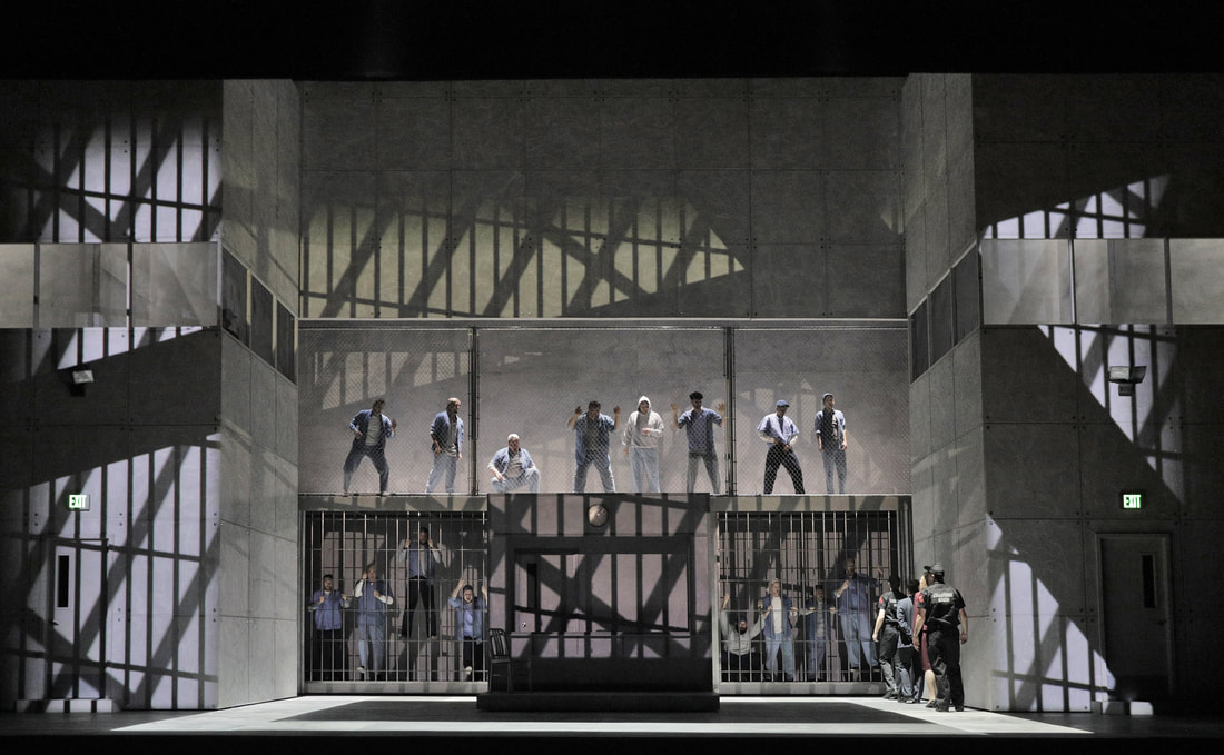 Picture: A drab set of squares and shadows of jail bars loom over the chorus, dressed as prisoners. 